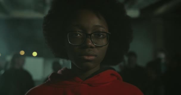 Vista da vicino della giovane ragazza afroamericana in occhiali che guarda alla telecamera mentre si trova alla manifestazione di notte. Una folla di manifestanti sullo sfondo. Concetto di sciopero . — Video Stock