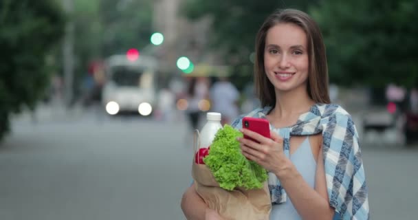 Mooie vrouw in casual kleding met behulp van smartphone tijdens het vasthouden van tas met voedsel. Millennial meisje scrollen nieuwsfeed en op zoek naar camera tijdens het staan op straat. Concept van winkelen. — Stockvideo