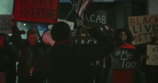 Le type au mégaphone se tournant vers la caméra et marchant tout en levant le poing serré et criant avec des manifestants derrière la foule. Grève contre la brutalité policière et le racisme la nuit. — Video