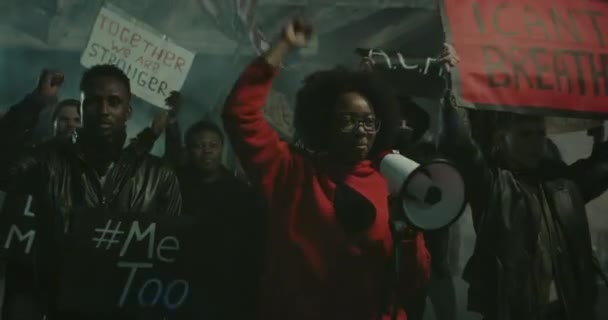 Chica joven con megáfono cantando y levantando el puño cerrado con la gente detrás. Multitud de manifestantes con pancarta de cartón y bandera americana caminando y gritando en la calle nocturna . — Vídeos de Stock