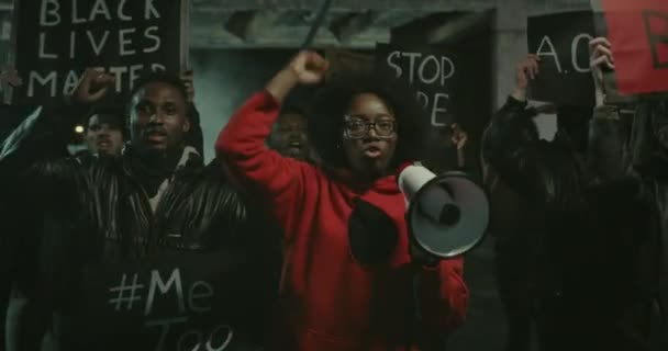 Chica afroamericana con megáfono cantando y levantando el puño apretado con la multitud detrás. Manifestantes emocionales con pancarta de cartón y bandera americana caminando y gritando en la calle . — Vídeos de Stock