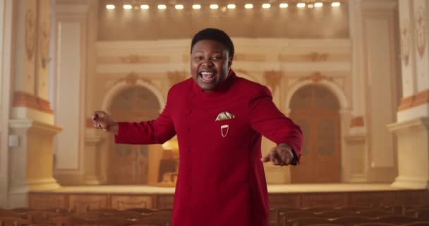 Crop view of charismatic musician in red suit looking to camera and singing while standing at row of wooden pews. Young guy performing gospel music and moving hands in rhytm. Zoom in. — Stock Video