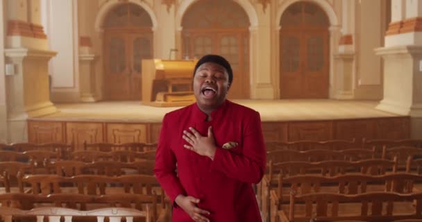 Artista afroamericano masculino con elegante traje rojo cantando mientras está de pie en la fila de bancos de madera en el gran salón. Joven talentoso interpretando música gospel emocionalmente y moviendo las manos . — Vídeo de stock