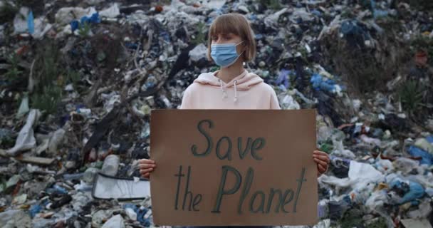 Mujer con máscara protectora girando la cabeza y mirando a la cámara mientras apoya guardar el movimiento de la tierra. Jovencita sosteniendo el estandarte de Save the Planet mientras está de pie frente a la colina de basura . — Vídeo de stock