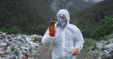 Koruyucu giysili bir adam ormanda çöp kovalarının yanında yürüyor. Solunum cihazı ve eldiven takmış plastik şişelere bakıp duran bir erkek. Çevre kirliliği kavramı.