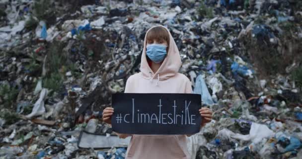 Girl in protective mask turning head and looking to camera while supporting eco awarness movement. Woman with hood holding climatestrike banner while standing near trash hill. Zoom in. — Stock Video