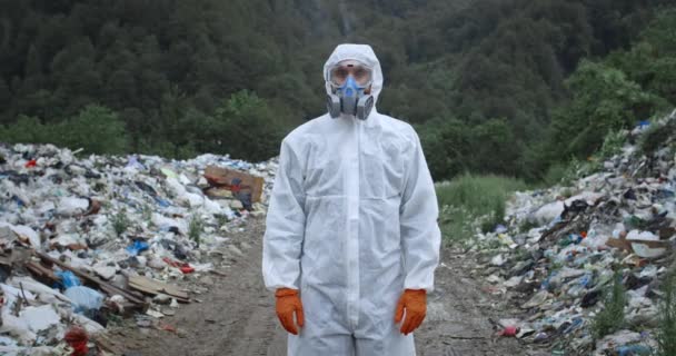 Männliche Person in Schutzkleidung steht unter Regen in der Nähe von Mülleimern im Wald. Mann mit Atemschutzmaske und Handschuhen blickt in die Kamera. Konzept von Umweltverschmutzung und Umweltbewusstsein. — Stockvideo