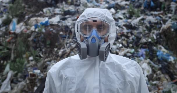 Vista de cerca de la persona masculina en el respirador de pie frente a una enorme colina de basura. Hombre con ropa protectora girando la cabeza y mirando a la cámara. Concepto de contaminación ambiental . — Vídeos de Stock