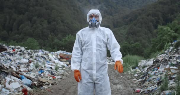 보호 복을 입은 남자 가 숲 속 쓰레기통 근처에서 비를맞고 서 있습니다. 인공호흡기를 끼고 장갑을 끼고 카메라를 찾고 정지 표시를 하는 남자. 환경 오염에 대한 개념. — 비디오