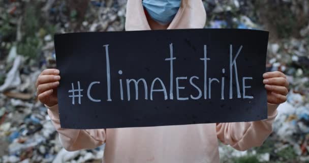 Primer plano de la mujer en máscara protectora levantando cartón negro con escritura huelga climática. Mujer en sudadera con capucha protestando contra la contaminación de la tierra mientras está parada frente a la colina de la basura . — Vídeo de stock