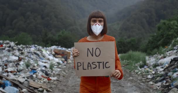 Crop view of girl in protective mask looking to camera while striking against pollution. Female activist holding no plastic banner while walkiing near trash pails in forest. — Stock Video