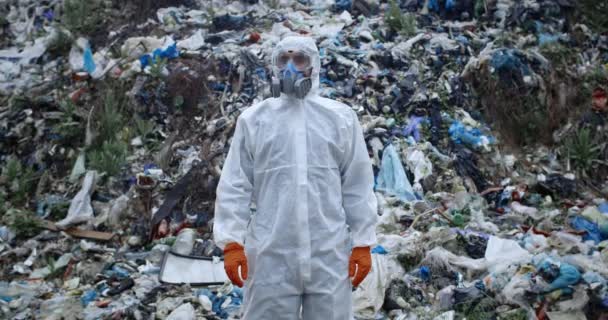 Чоловік у захисному костюмі та респіраторі стоїть перед величезним сміттєвим пагорбом. Людина в захисному одязі повертає голову і дивиться на камеру. Концепція забруднення навколишнього середовища . — стокове відео
