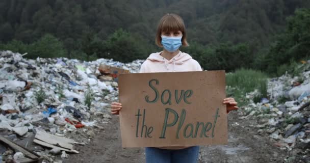 Дівчинка в захисній масці дивиться на камеру, підтримуючи рух Землі. Молода жінка, що тримає прапор "Врятуйте планету", коли йде під дощем біля сміттєзвалища. Концепція екології. — стокове відео