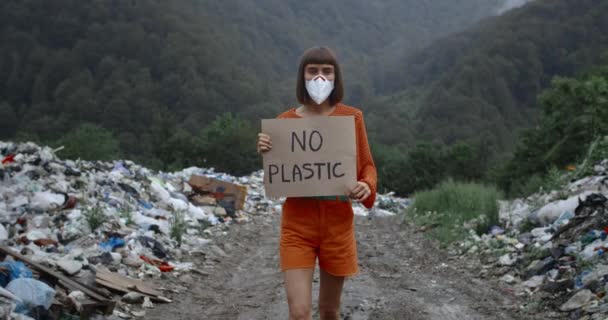 Menina bonita em máscara protetora em seu rosto olhando para a câmera enquanto golpeia contra a poluição. Mulher ativista segurando nenhuma caixa de plástico enquanto passeia perto de pails lixo na floresta . — Vídeo de Stock