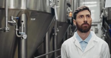 Beyaz laboratuvar önlüklü yakışıklı adam yürüyor ve yana bakıyor. Kendine güvenen erkek işçi fabrikadaki otomatik içecek makinesini kontrol ediyor. Bira fabrikası ve bira fabrikası kavramı.