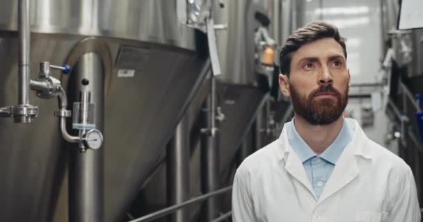 Hombre guapo y concentrado en bata blanca caminando y mirando a un lado. Trabajador masculino seguro que comprueba el trabajo de la máquina automática de la bebida en fábrica. Concepto de fábrica de cerveza y planta de cerveza . — Vídeos de Stock