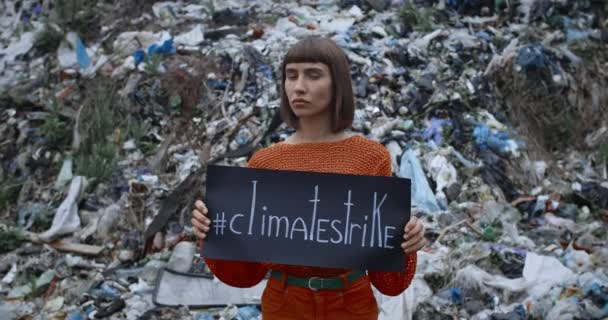 Millennial flicka vrida huvudet och titta på kameran samtidigt protesterar mot miljöföroreningar. Olycklig ung flicka som håller klimat strejk tecken när du står nära skräp kulle. — Stockvideo
