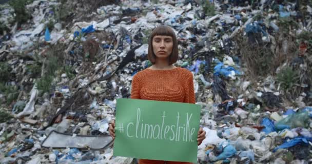 Mujer joven con un anillo en la nariz levantando un cartel verde con el hashtag de la huelga climática mientras está de pie frente a la colina de basura. Concepto de protección y contaminación ecológicas. Acercar . — Vídeos de Stock