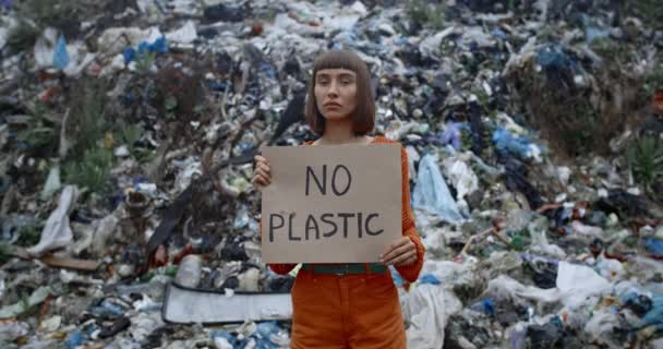 Dívka s kroužkem v nose drží karton bez plastového nápisu, zatímco stojí před odpadkovým kopcem pod deštěm. Žena otáčí hlavu a dívá se do kamery, zatímco protestuje proti znečištění. — Stock video
