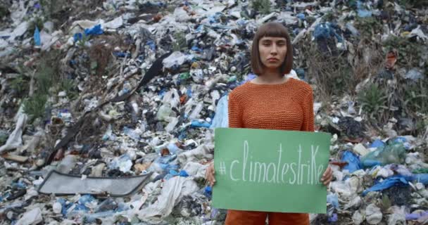 Eine ernsthafte junge Frau hält vor einem Müllberg eine grüne Plakette mit der Aufschrift "Klimawürger" in der Hand. Kornblick eines schönen Mädchens, das gegen Umweltverschmutzung protestiert. — Stockvideo
