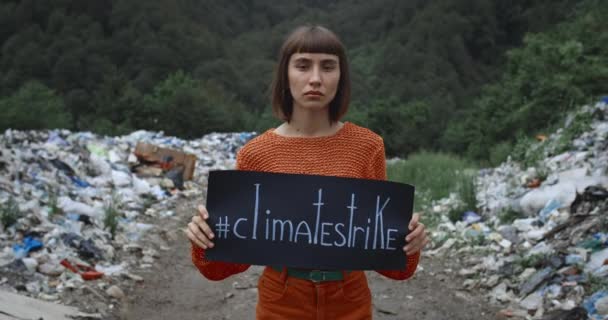 Chica joven seria sosteniendo pancarta con climatestrike escribir en ella mientras camina cerca de pilas de basura. Vista de la cosecha de la mujer milenaria protestando contra la contaminación ambiental . — Vídeo de stock