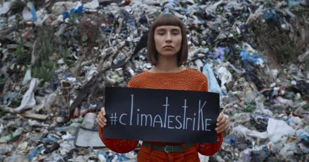 Woman with nose ring turning head to camera while holding poster with climate strike hashtag. Serious girl standing in front of trash hill. Concept of ecology protection and pollution. — Stock Video