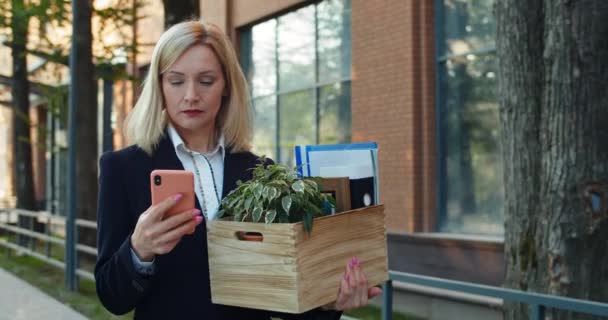 Verontrustende vrouw in de 30 in een formeel pak met haar spullen op straat. Vrouwelijke werknemer lopen en scrollen smartphone scherm tijdens het zoeken naar nieuws. Begrip werkloosheid. — Stockvideo