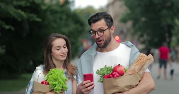 Happy couple using smartphone and saying wow while having good news at smartphone . Girl and guy coming back after shopping and carrying food in paper bags while walking at city street. — Stock Video