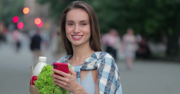 Csinos lány portréja, amint a közösségi oldalakon cseveg, és az utcán áll, miközben kamerára mereszti a szemét. Egy nő okostelefont használ, miközben zacskót tart étellel. A vásárlás fogalma. — Stock videók