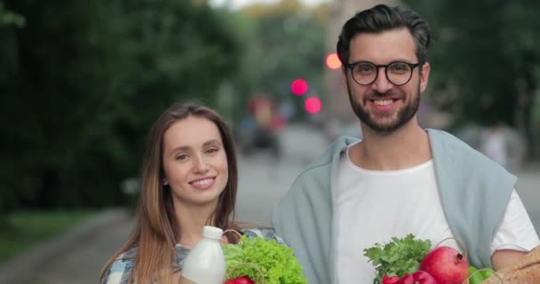 Close up view of cheerful young family looking to each other and to camera . Crop view of smiling man and woman holding craft paper bags with food and standing at street. — Stock Video