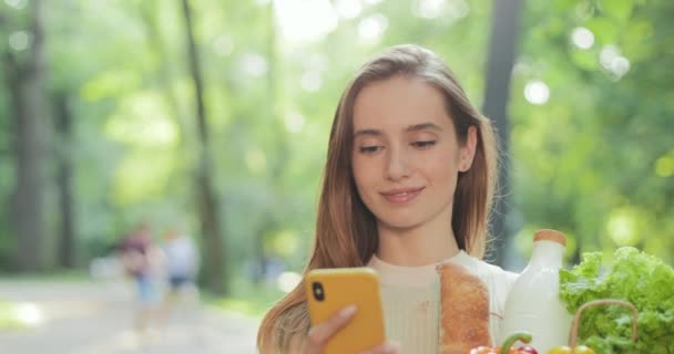 Hübsche Frau, die Nachrichten checkt und lächelt, während sie ihr Smartphone benutzt. Millennial Girl trägt Lebensmittel in einer Tasche, während sie geht und auf den Bildschirm schaut. Reflexionen des Sonnenlichts. Unklarer Hintergrund. — Stockvideo