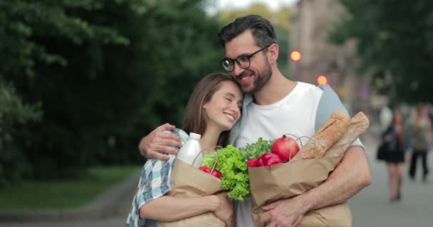 歩いている間に甘いカップルがお互いを見て楽しい時間を過ごしているのを間近で見ることができます。路上で食べ物と紙の袋を持っている間、彼らは彼の美しい妻を抱きしめる眼鏡の髭の男. — ストック動画