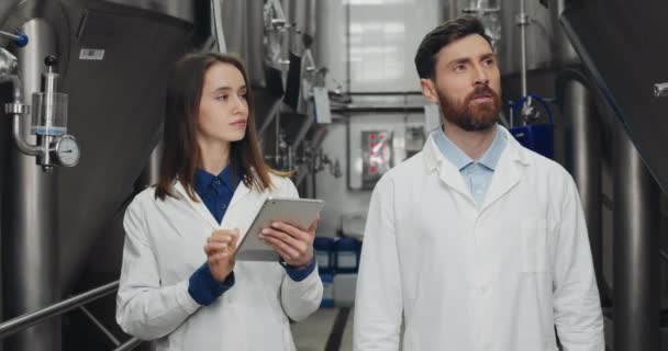 穿着白衣的男工和女工检查设备。男人在口述数据，女人在平板电脑上输入数据时，一边走着一边翻看一排排的炼钢桶. — 图库视频影像
