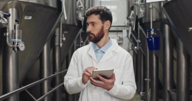 Sakallı erkek işlem mühendisi bira fabrikasındaki tablete veri giriyor. Beyaz laboratuvar önlüklü yakışıklı adam yürürken ve ekipman göstergelerine bakarken dijital ekrana dokunup yazıyor..