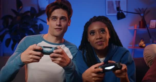 Happy young people playing video games on console while sitting on couch in front of tv. Millennial couple spending fun time together at home.Room with warm and neon lights. — Stock Video