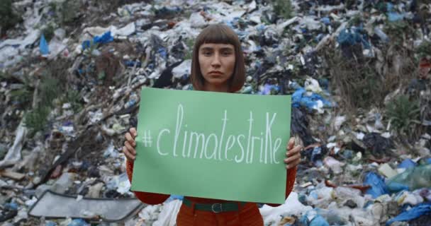 Hermosa chica sosteniendo cartel verde con etiqueta de huelga climática mientras está de pie frente a la colina de la basura. Mujer milenaria en huelga por la ecología y el planeta limpio. Concepto de salvar la Tierra . — Vídeos de Stock