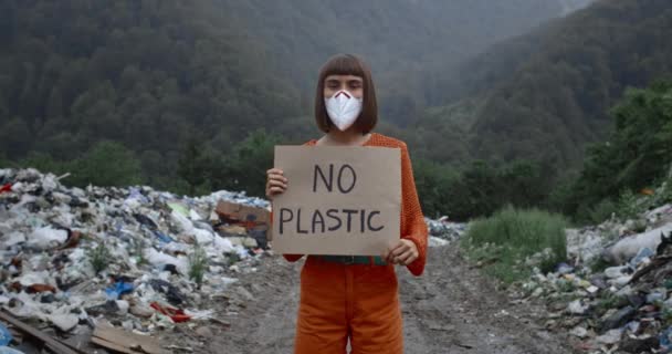 Mujer milenaria en máscara protectora girando cabeza y mirando a la cámara mientras golpea contra la contaminación. Chica sosteniendo ninguna pancarta de plástico mientras está de pie cerca de cubos de basura en el bosque . — Vídeos de Stock