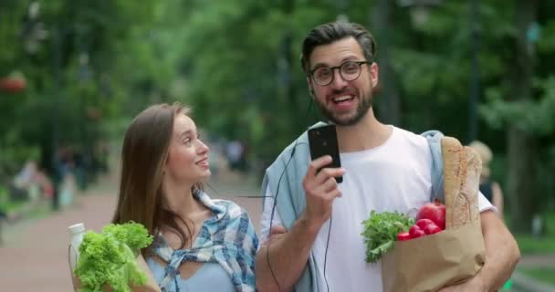 Pareja escuchando música juntos en auriculares y cantando en voz alta mientras caminan en el parque. Hombre y mujer pasándolo bien mientras llevan la bolsa con comida. Concepto de relación feliz . — Vídeo de stock