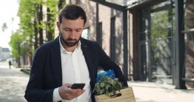 30 'lu yaşlarda üzgün bir adam resmi takım elbise giyiyor ve sokakta eşyalarıyla birlikte kutu taşıyor. Haberleri ararken erkek çalışan yürür ve akıllı telefon ekranını kaydırır. İşsizlik kavramı.