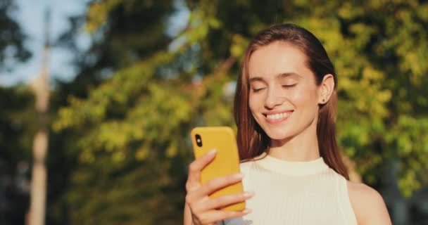 Close-up zicht op mooie brunnette vrouw op zoek naar camera en glimlachen tijdens het gebruik van de smartphone. Gelukkig meisje scrollende telefoon scherm terwijl buiten. wazig park achtergrond. — Stockvideo