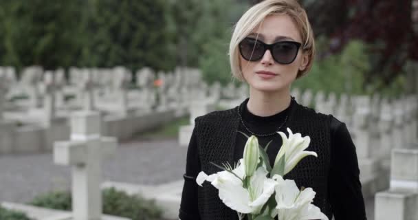 Großaufnahme einer Frau mit dunkler Brille, die den Kopf hebt und in die Kamera blickt. Junge Witwe mit weißer Lilienblüte auf dem Friedhof. Konzept des Gedenktages. Unklarer Hintergrund. — Stockvideo