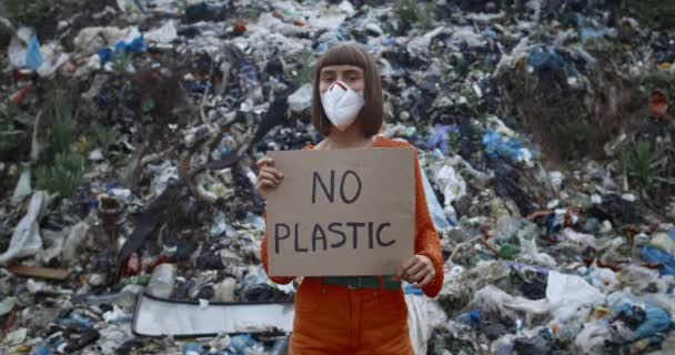 Meisje met een beschermend masker die karton vasthoudt zonder plastic schrift terwijl ze voor de vuilnisheuvel staat onder de regen. Draaiende vrouw die naar de camera kijkt terwijl ze protesteert tegen vervuiling. — Stockvideo
