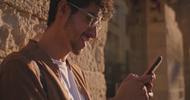 Şehir caddesinde duvara yaslanmış beklerken gözlüklü adamın telefonla gülüşünü yakından çek. Sosyal ağlarda iletişim kurarken burun halkalı neşeli adam mesaj yazıyor..