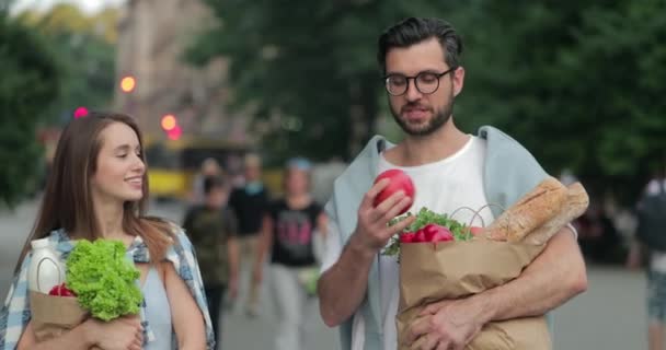 快乐的夫妇拿着装满食物和谈话的纸袋。戴眼镜的大胡子男人一边和他漂亮的妻子散步一边扔石榴。混淆不清的街道背景. — 图库视频影像