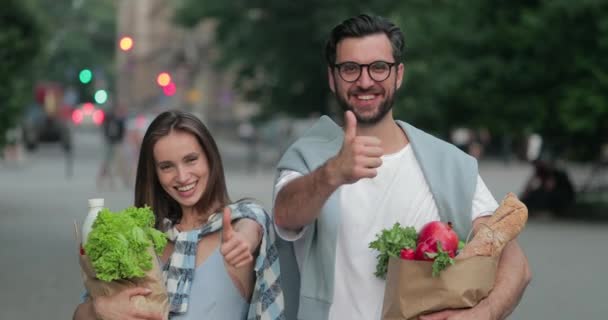 Retrato de pareja alegre sonriendo y mostrando el pulgar hacia arriba mientras está de pie en la calle. Vista de la cosecha del hombre en gafas y su novia sosteniendo bolsas de papel artesanales con comida saludable. Acercar . — Vídeos de Stock