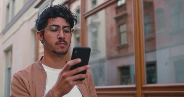 Crop view van de jonge man met de baard in glazen met behulp van zijn smartphone tijdens het lopen op straat. Guy met neusring op zoek naar telefoonscherm tijdens het surfen op internet in de stad straat. — Stockvideo