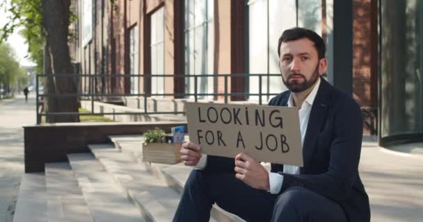 Unhappy young man showing looking for job carton banner near office building. Dismissed male worker in suit searching for work while sitting on stairs. Concept of unemployment. — Stock Video