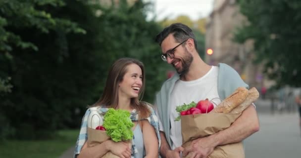 Hombre alegre en vasos arrojando granada mientras habla y camina con su atractiva esposa.Pareja joven llevando bolsas de papel llenas de comida. Fondo de calle borrosa . — Vídeos de Stock