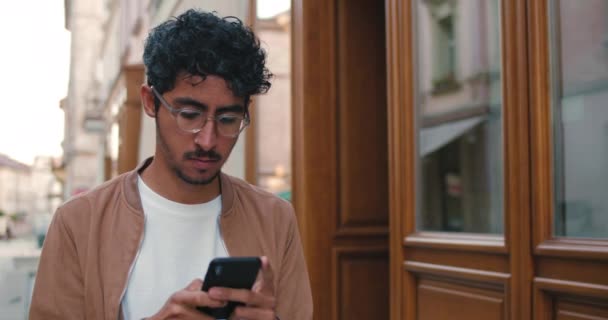 Millennial 'daki gözlüklü adam sokakta yürürken akıllı telefonunu kullanıyor. Sokakta sosyal ağlarda sohbet ederken daktilo eden ve telefon ekranına bakan yakışıklı bir adam.. — Stok video