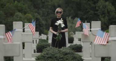 Mezarlıkta elinde beyaz zambak çiçeği tutan kadının portresi. Siyah gözlüklü dul, Amerikan bayraklarıyla sıralanmış taş haçlar arasında dikilirken kameraya bakıyor. Anma Günü kavramı.